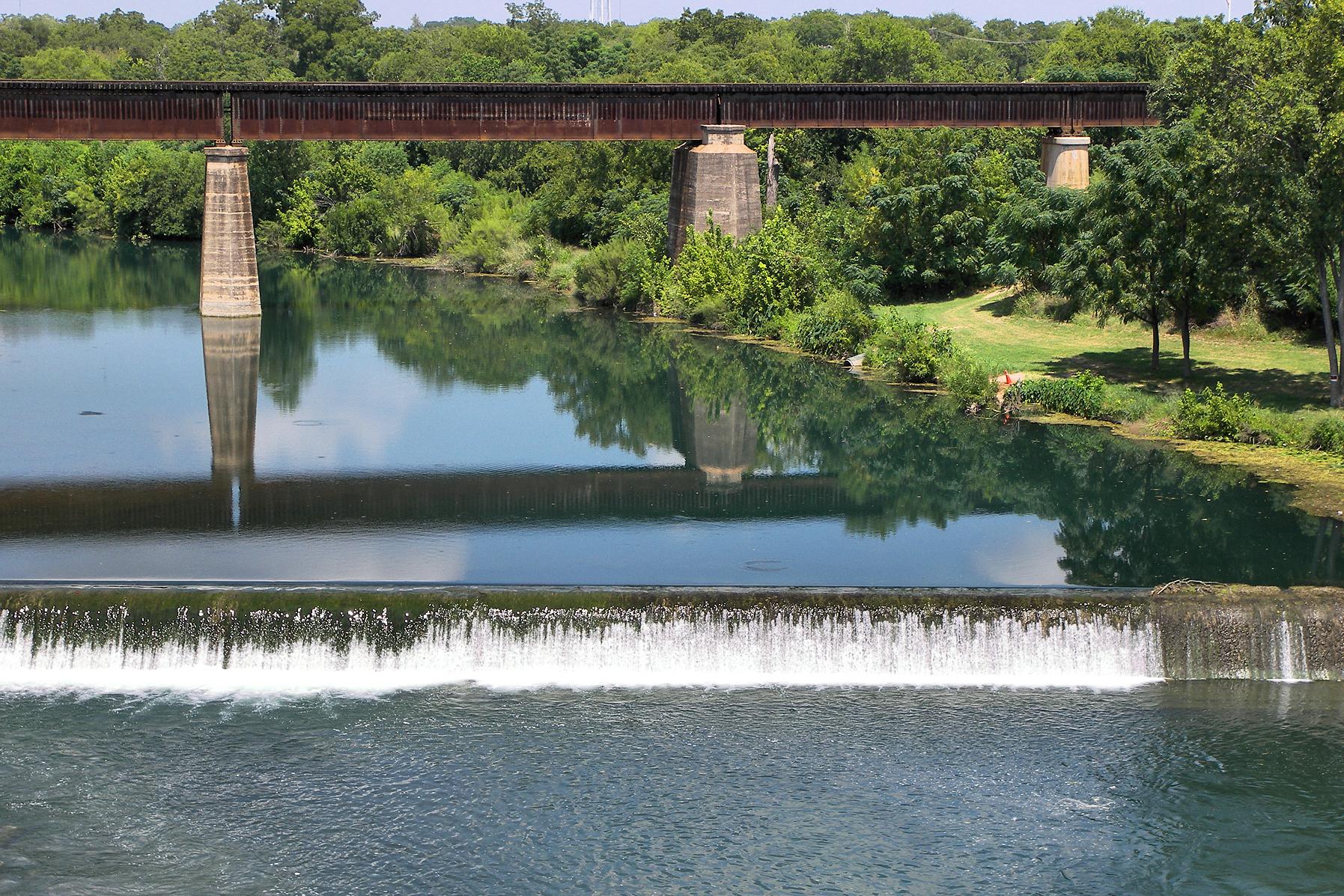 A low dam spilling across the Guadalupe River with an old railroad truss bridge behind