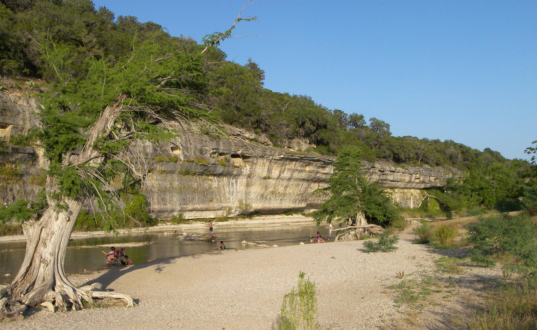 A limestone bluff and cypress trees along the Guadalupe River, with people wading by a gravel beach