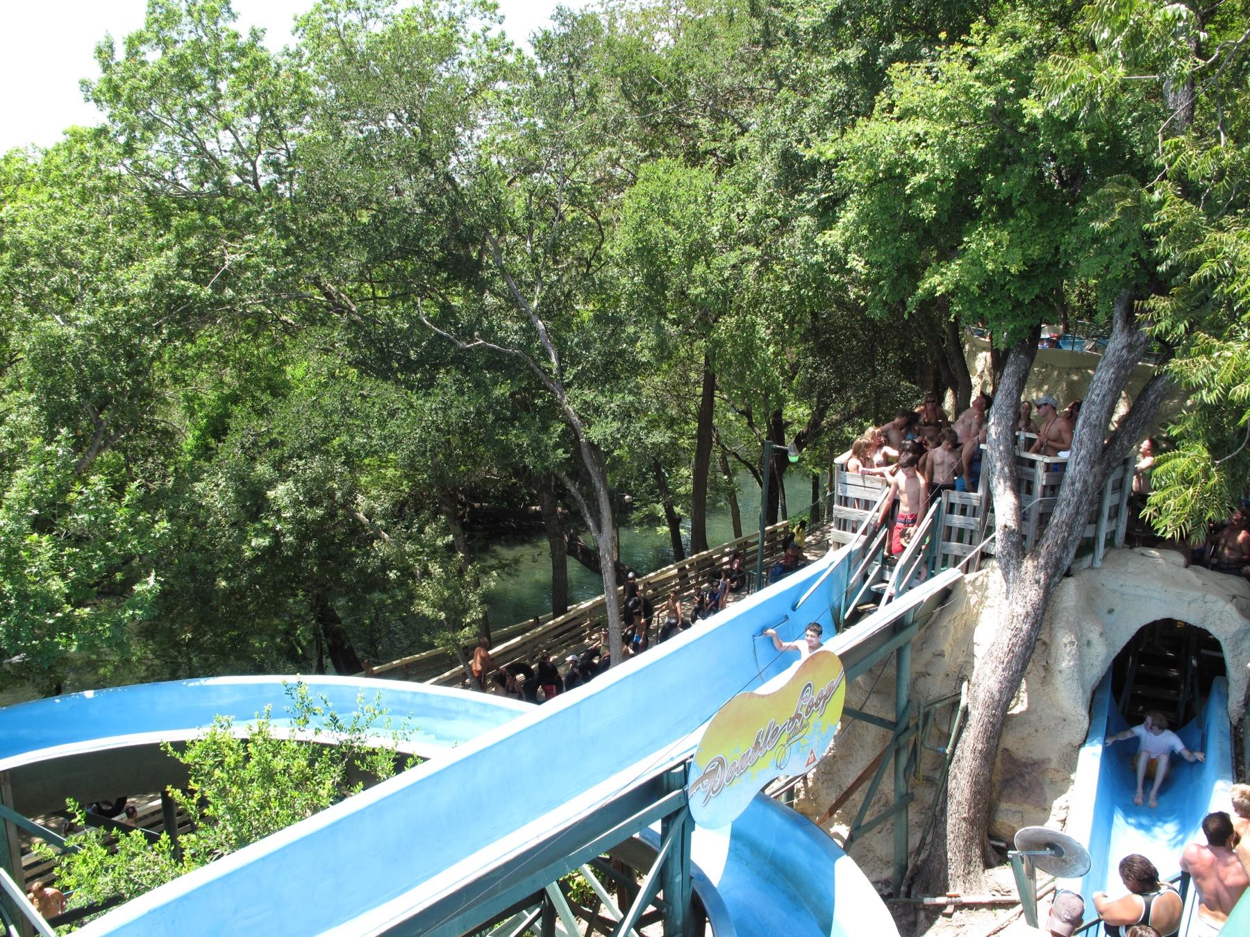 A blue water slide winding under shade oaks at Schlitterbahn, riders queuing on the wooden platform