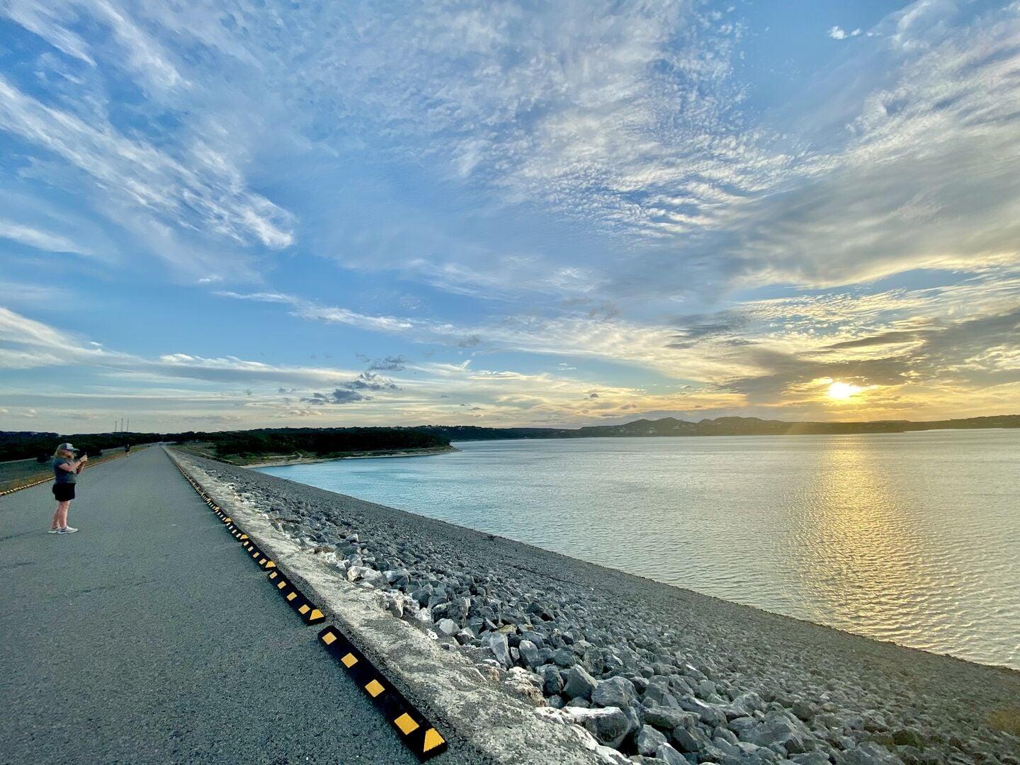 A person standing on the paved crest of Canyon Dam at sunset, the lake stretching to the horizon